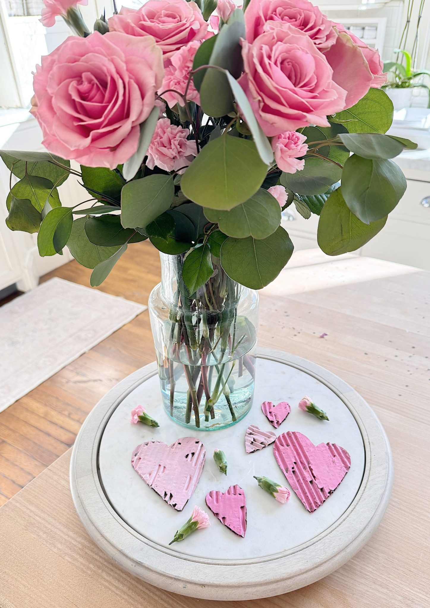 A glass vase with pink roses and greenery stands on a round marble tray, next to pink heart-shaped decorations and small pink flower buds.