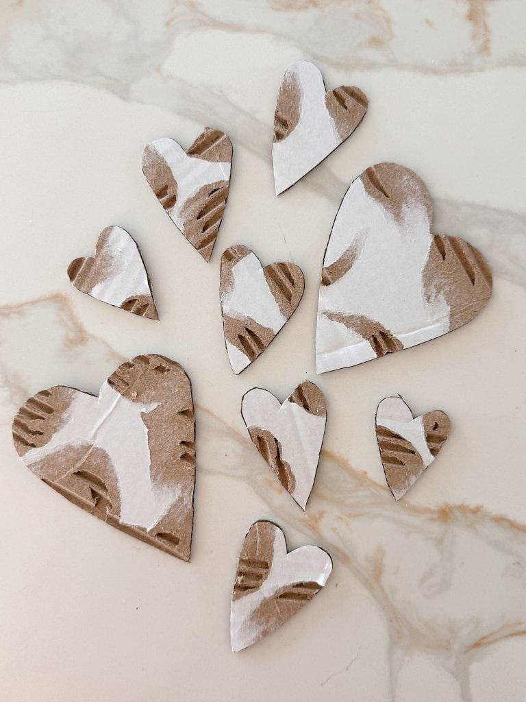 Nine heart shapes cut from brown and white corrugated cardboard are arranged on a light marble surface.