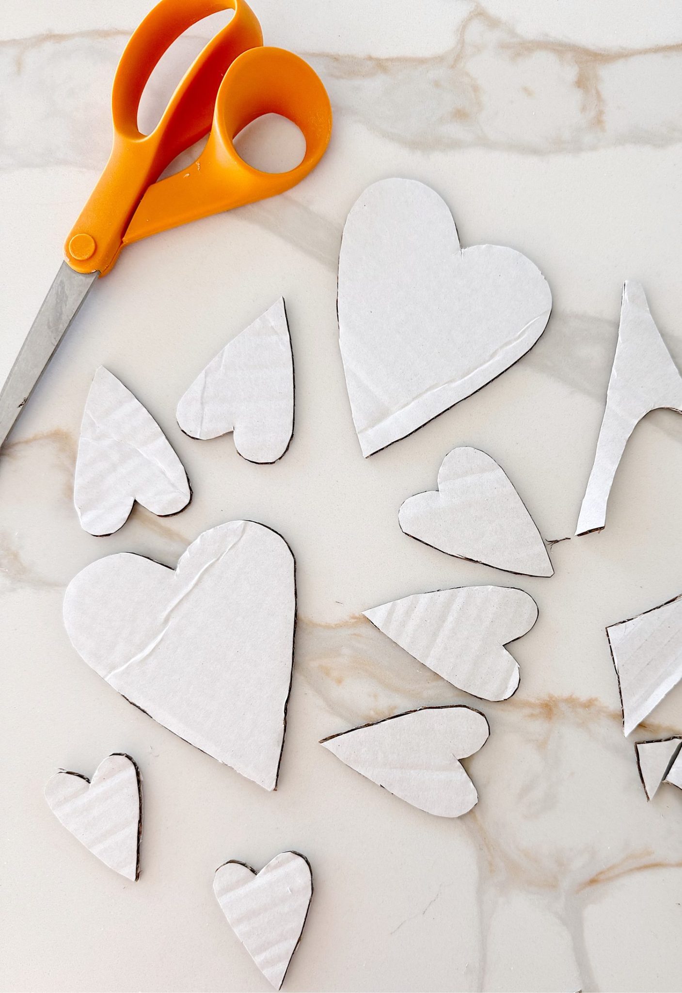 Orange scissors and several white cardboard hearts of various sizes are placed on a marble surface, with some heart shapes yet to be fully cut out.