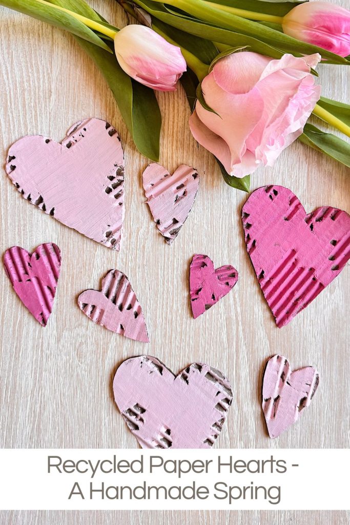 Pink and purple recycled paper hearts displayed on a wood surface, with a bouquet of pink tulips and roses above. Text reads: "Recycled Paper Hearts - A Handmade Spring.
