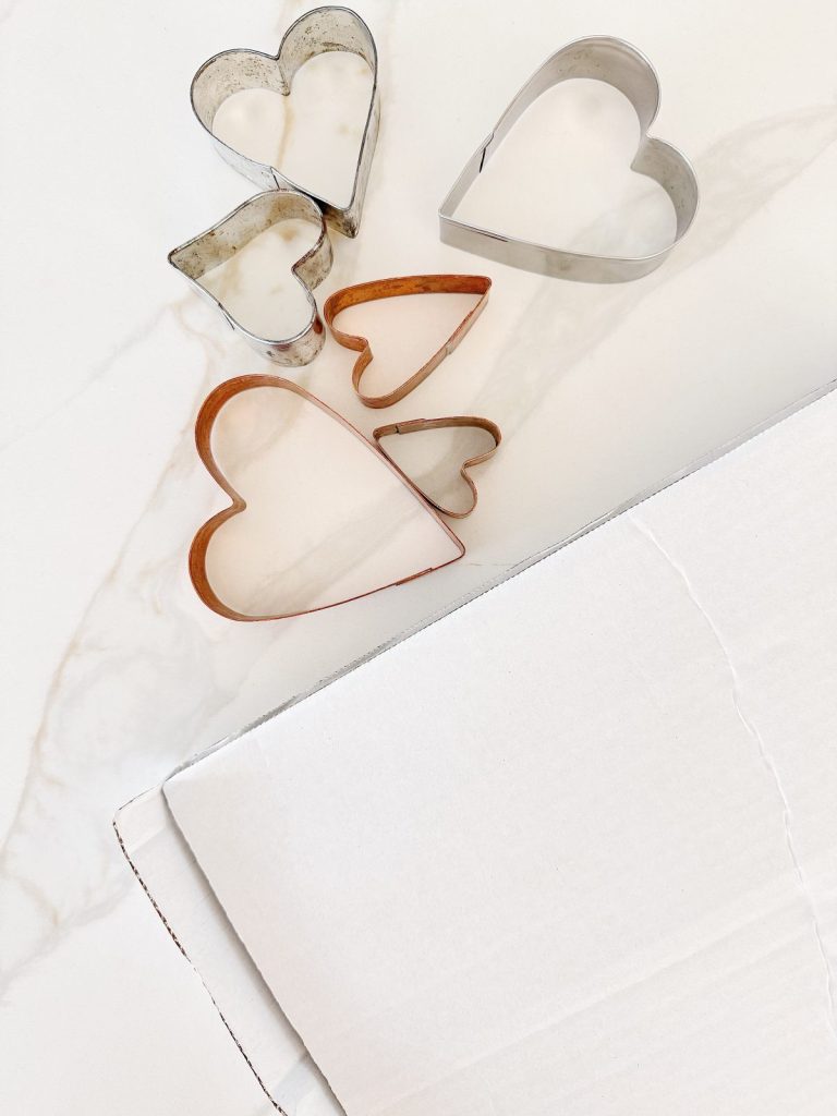 Five heart-shaped cookie cutters of various sizes rest on a white marble surface next to the edge of a large white cardboard box.