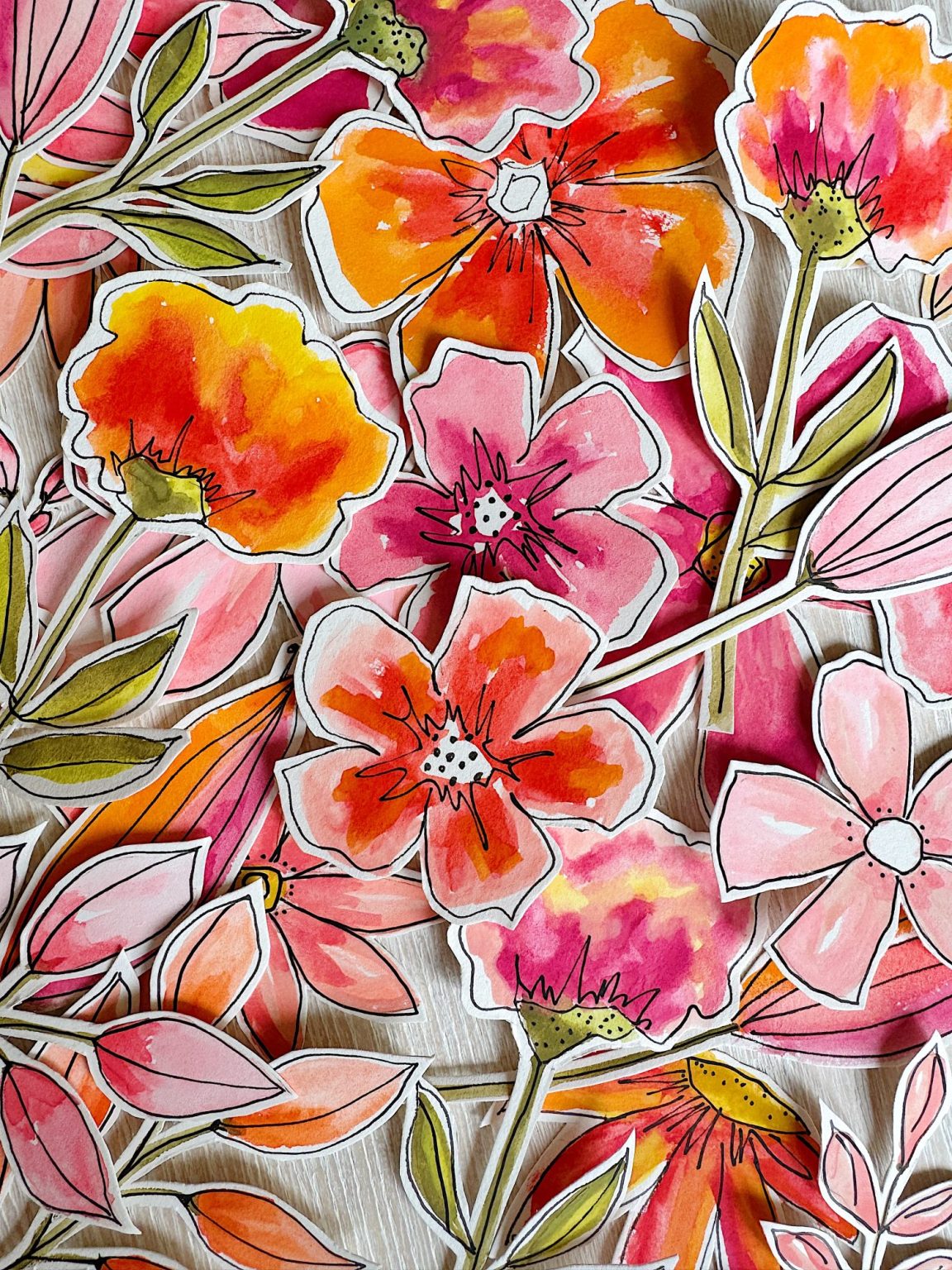 Hand-drawn and watercolor-painted paper flowers in shades of pink, orange, and red are scattered on a light wooden surface.