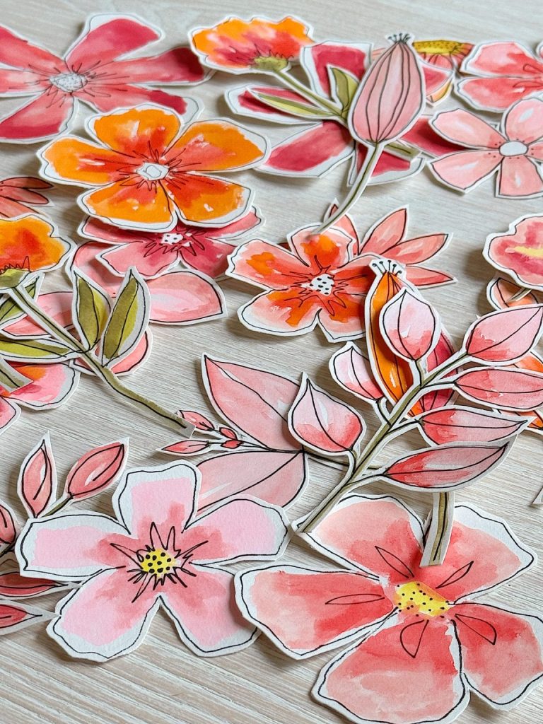 Cut-out watercolor illustrations of pink, orange, and red flowers with green leaves scattered on a light wooden surface.