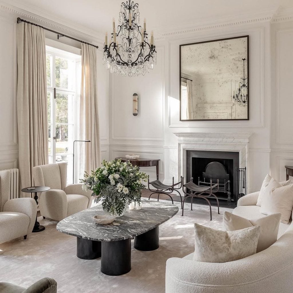 Elegant living room with cream-colored furniture, a marble coffee table, large window, ornate chandelier, fireplace, and a large mirror above the mantel.