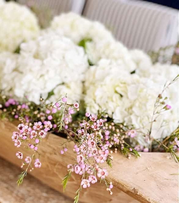 A wooden container holds a floral arrangement with white hydrangeas and small pink flowers, set against a blurred background with striped cushions.