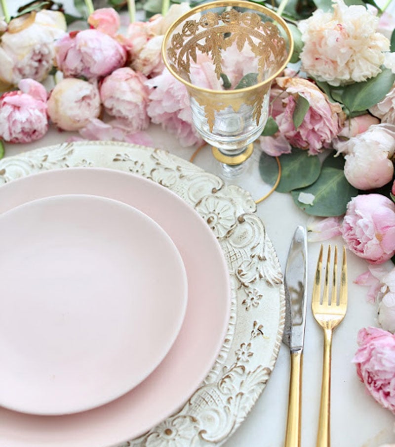 A table setting with pink plates on a decorative white charger, gold cutlery, a gold-rimmed glass, and pink peonies in the background.