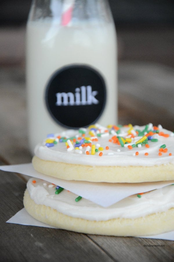 Two frosted sugar cookies with colorful sprinkles are stacked on parchment paper in front of a glass bottle of milk on a wooden surface.