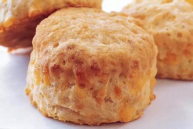 Close-up of a golden-brown biscuit with a flaky texture, featuring visible bits of cheese, resting on a white surface.