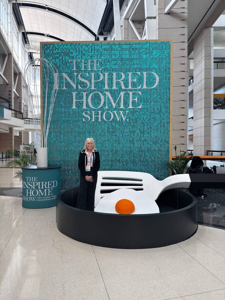 A woman stands in front of a large "The Inspired Home Show" sign, next to a giant egg sculpture with a black base, inside a spacious convention center.