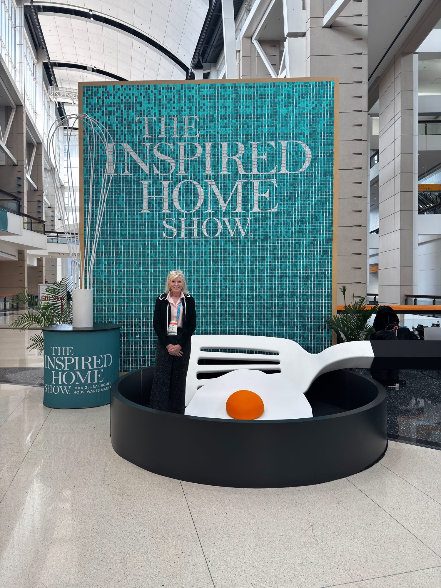 A woman stands in front of a large "The Inspired Home Show" sign, next to a giant egg sculpture with a black base, inside a spacious convention center.