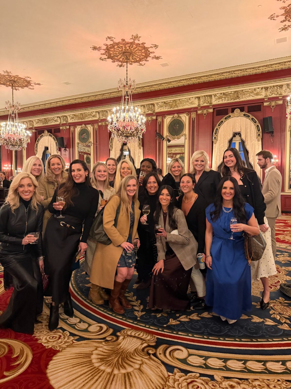 A group of people pose for a photo in an ornate, chandelier-lit room with red walls and gold accents. Many hold drinks and are dressed in semi-formal attire.