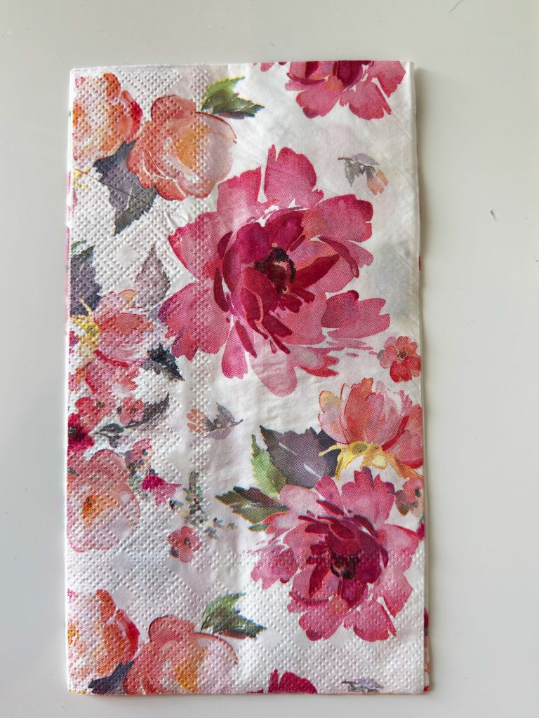 A folded white paper napkin with a pattern of pink and red flowers and green leaves.