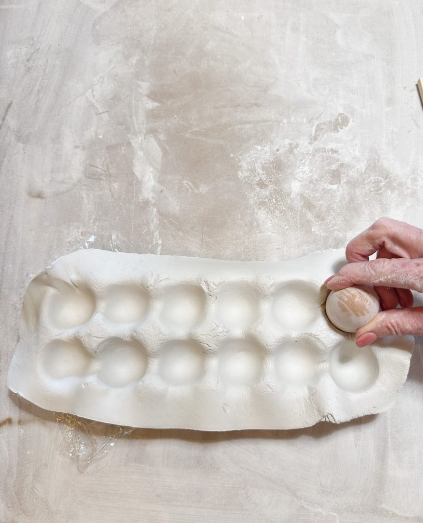 A hand presses a small round object into a white clay slab with twelve circular indentations on a light-colored surface.