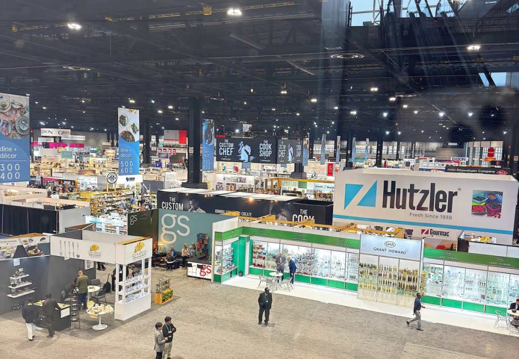 Large indoor trade show featuring inspired home company booths, banners, and displays; a few people are walking and browsing the exhibits.