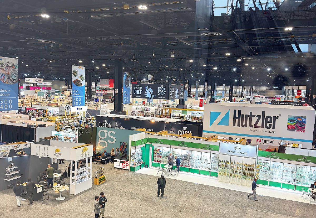 Large indoor trade show featuring inspired home company booths, banners, and displays; a few people are walking and browsing the exhibits.