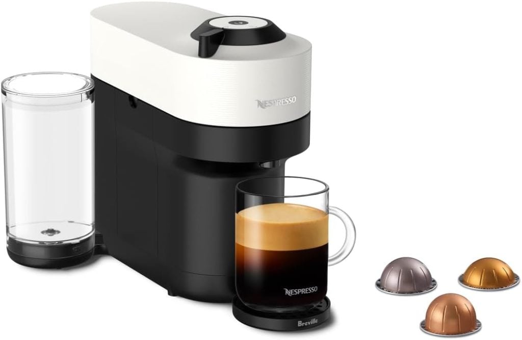A Nespresso coffee machine with a glass mug of coffee on its tray, a water reservoir at the back, and three coffee capsules nearby&mdash;perfect for upgrading your mornings during the Amazon Big Spring Sale.
