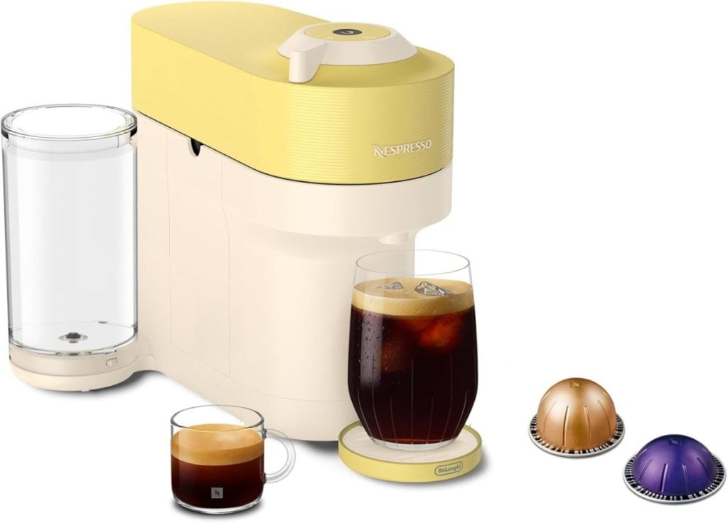 Yellow and white Nespresso coffee machine with a glass of iced coffee, a cup of espresso, three coffee pods, and a transparent water tank&mdash;perfect for your home brew setup this Amazon Big Spring Sale.
