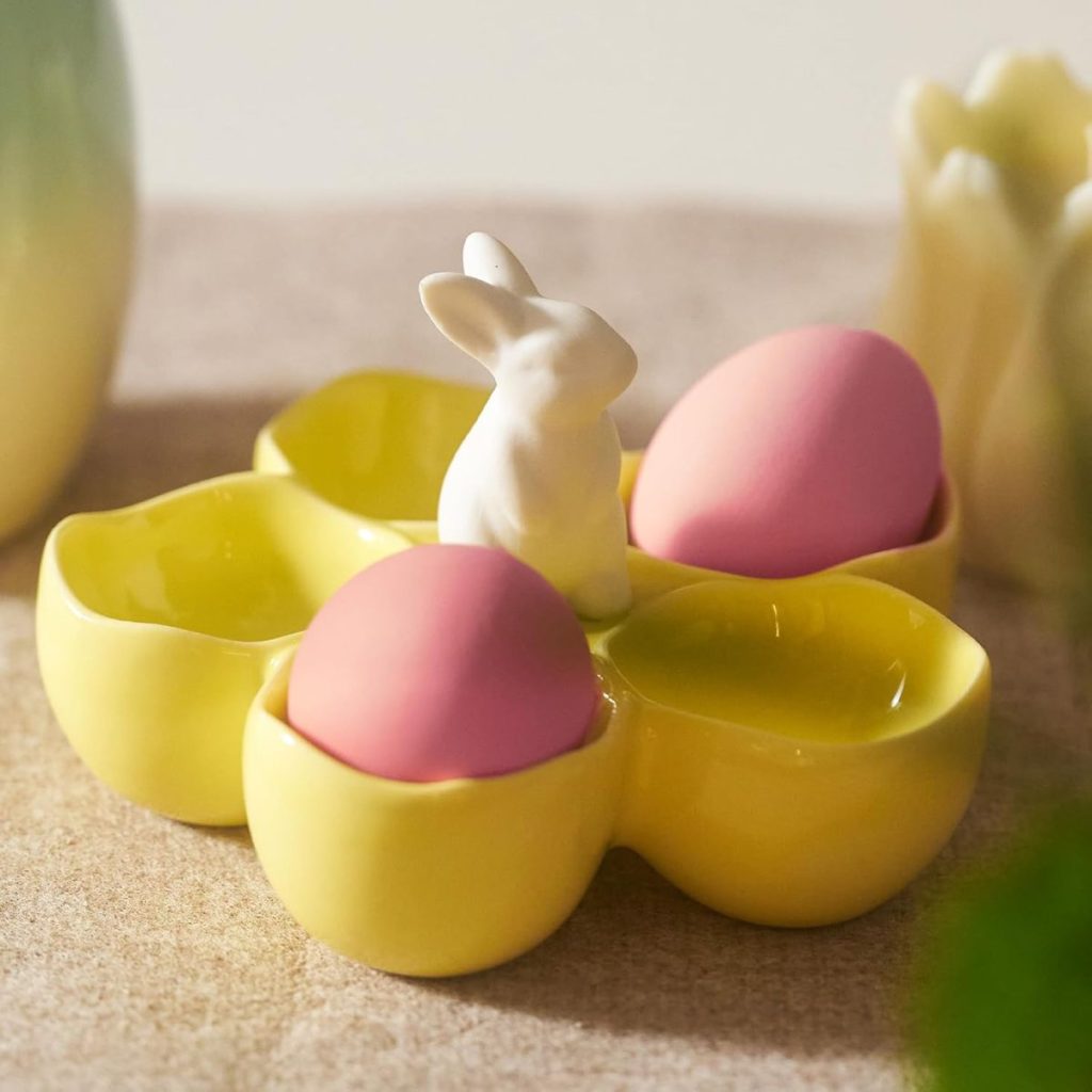 A yellow ceramic egg holder with a white rabbit figurine in the center holds two pink eggs.