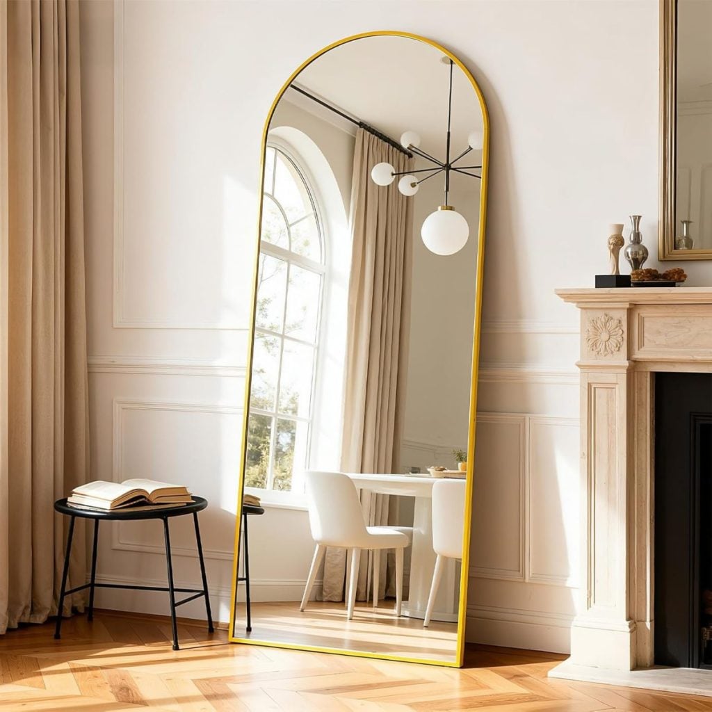 A large arched mirror with a gold frame stands against a wall, reflecting a dining area with a round table, white chairs, and a modern chandelier&mdash;an elegant look perfect for finds from the Amazon Big Spring Sale.