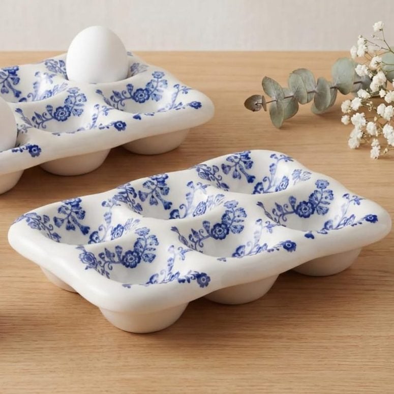 Two ceramic egg trays with blue floral patterns hold white eggs on a wooden surface, with extra eggs, a sprig of eucalyptus, and baby&rsquo;s breath nearby.