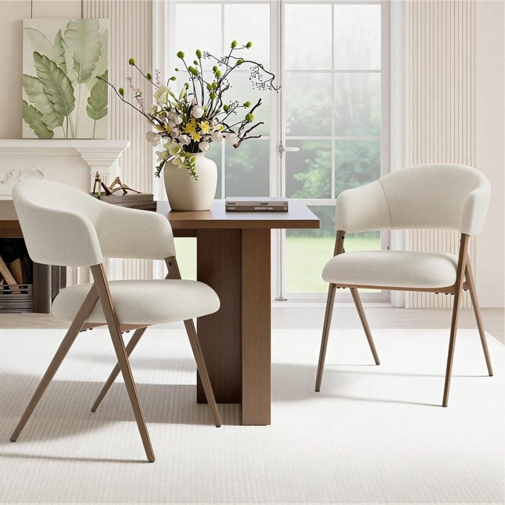 A modern dining area with two white cushioned chairs, a wooden table, and a vase of flowers, set in front of large windows with sheer curtains&mdash;perfect inspiration for your next Amazon Big Spring Sale find.