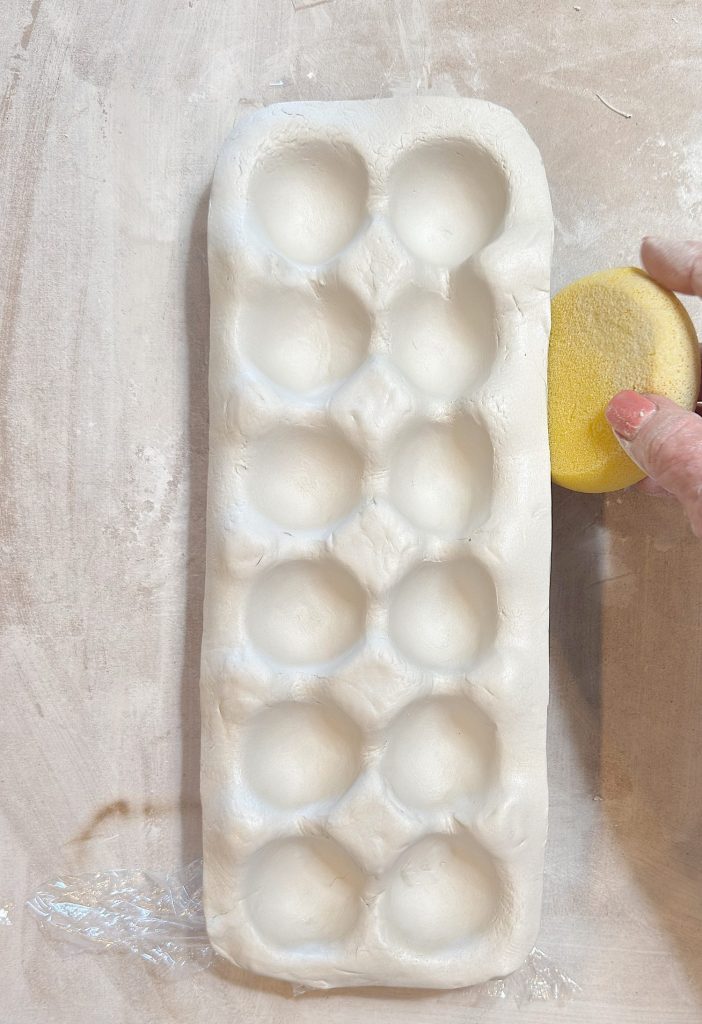 A hand smooths a white clay tray with round indentations using a yellow sponge on a light surface.