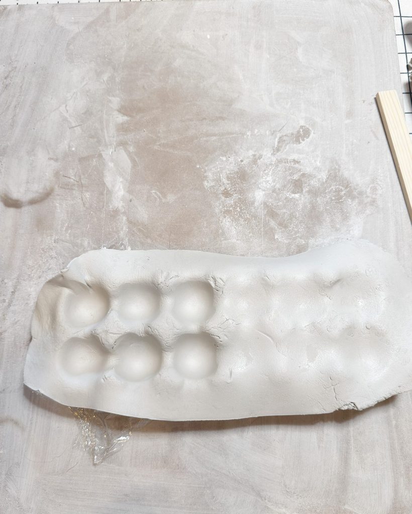 A slab of white clay with several rounded indentations lies on a floured surface next to a wooden stick.