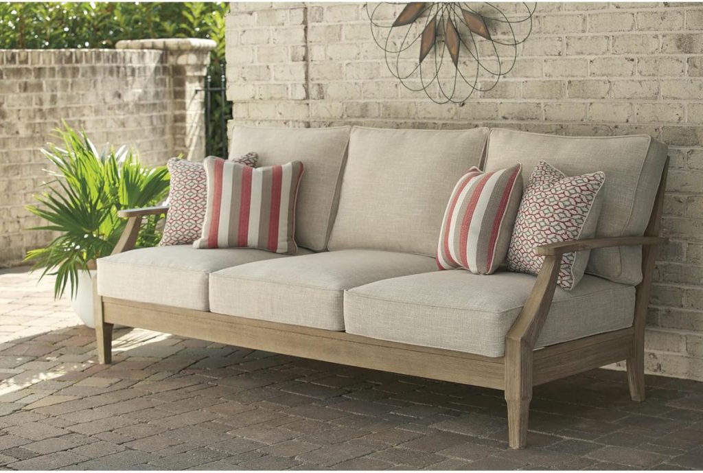 Outdoor beige cushioned sofa with wooden frame, three striped and patterned accent pillows, set on a brick patio beside a green plant and a light-colored brick wall&mdash;perfect for relaxing during the Amazon Big Spring Sale.