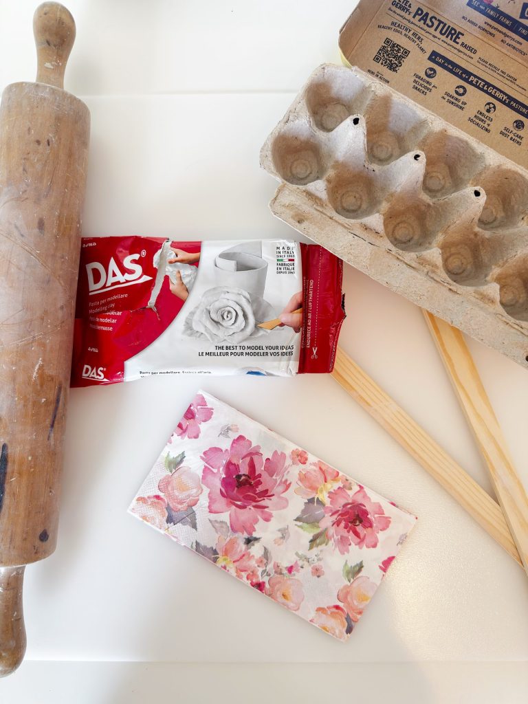 A rolling pin, a red pack of modeling clay, an empty egg carton, two wooden sticks, and a floral-patterned wallet are arranged on a white surface.