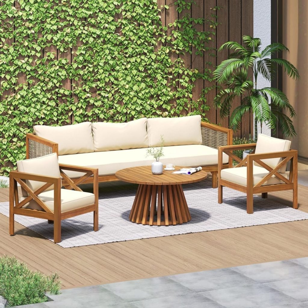 Elevate your outdoor space for the Amazon Big Spring Sale with this patio set: a wooden sofa, two armchairs with cream cushions, a round wooden table, and a potted plant on a rug, all surrounded by lush greenery and a wooden fence.