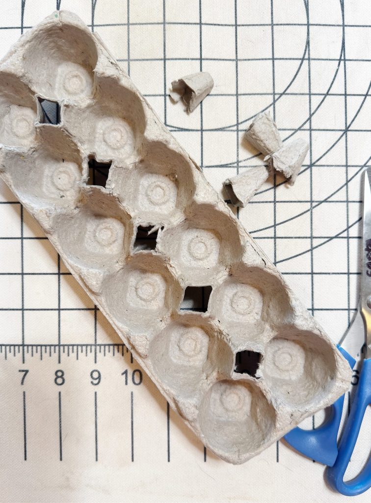 An egg carton with four square holes sits on a gridded surface next to cut-out carton pieces and a pair of scissors.