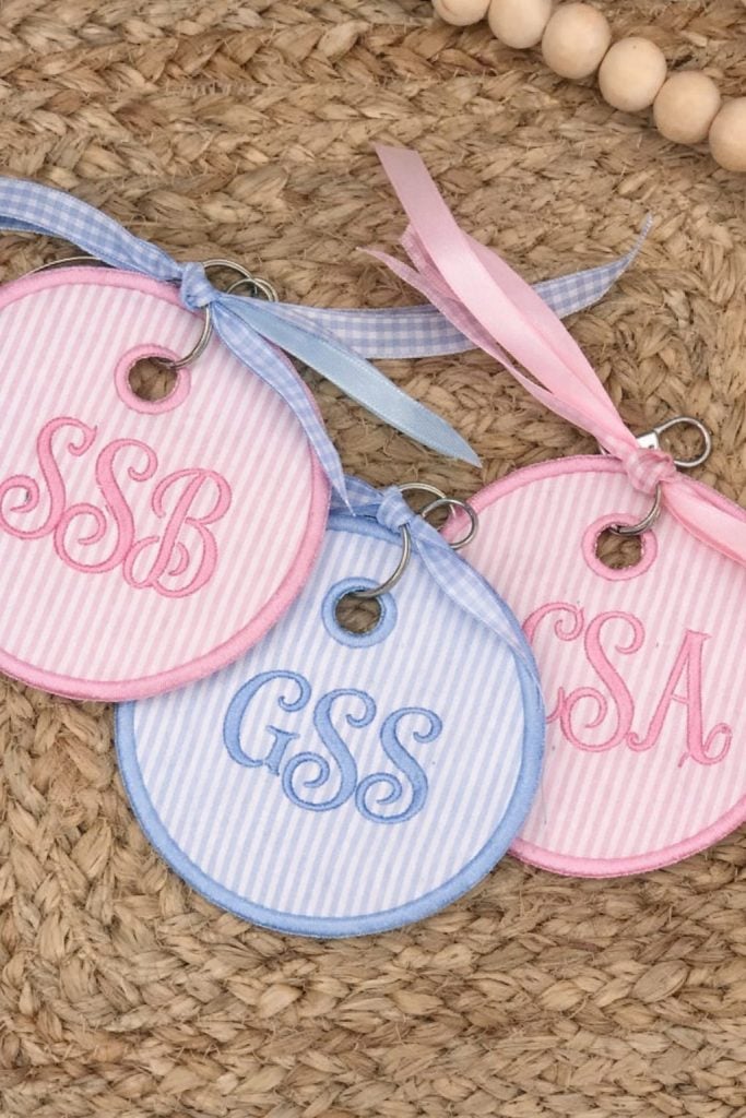 Three round, fabric keychains with embroidered initials (SSB, GSS, CSA) in pink and blue, each with a matching ribbon, are displayed on a woven surface.