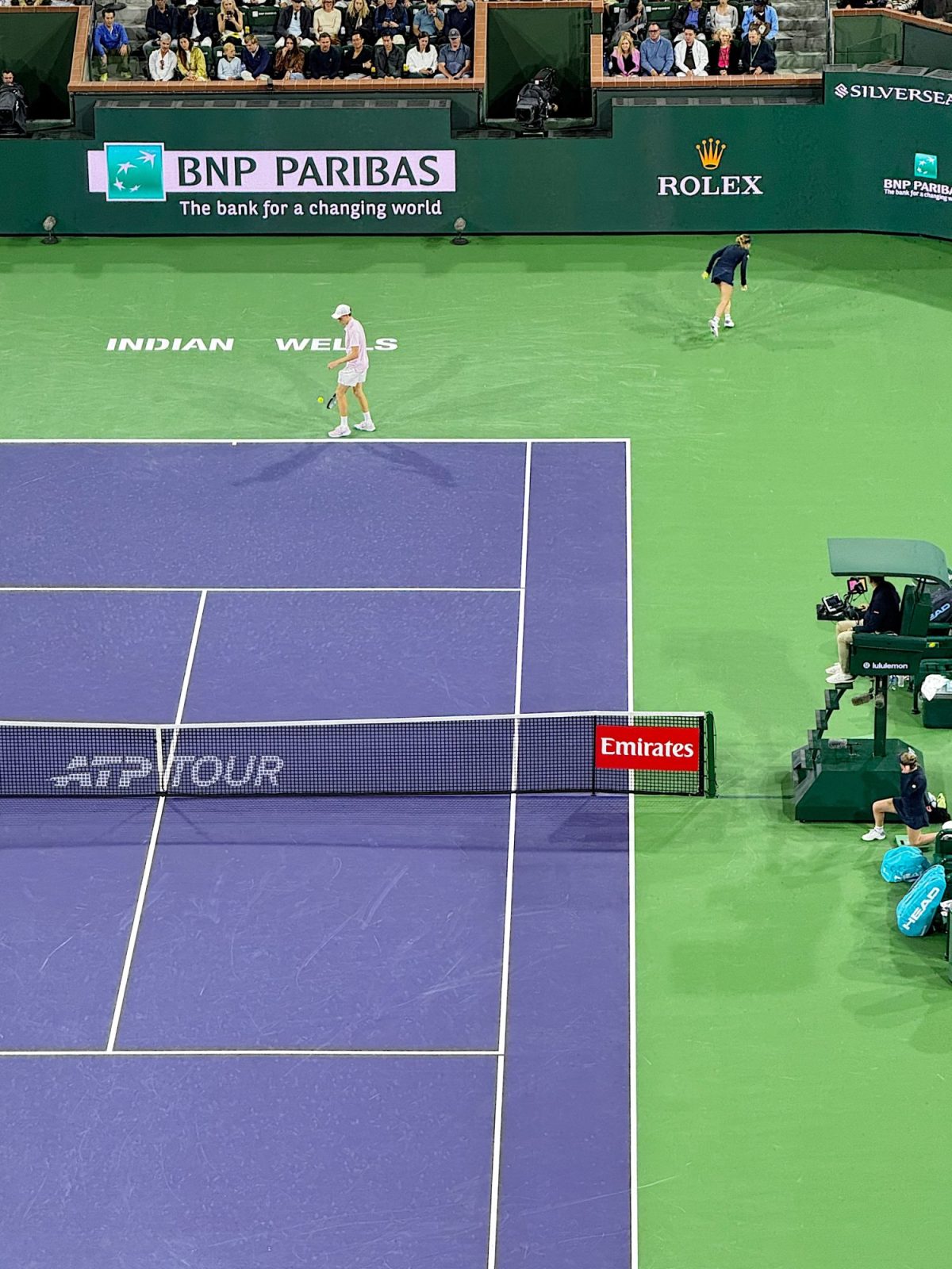 Two tennis players are on a hard court at Indian Wells during a match, with spectators watching and sponsorship banners visible around the court.