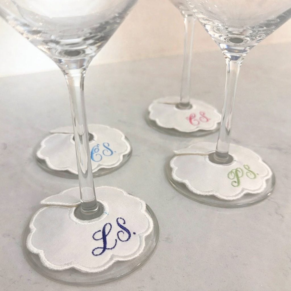 Four wine glasses with white, scalloped fabric tags on the stems, each embroidered with different colored initials: LS, EC, ES, and PS.