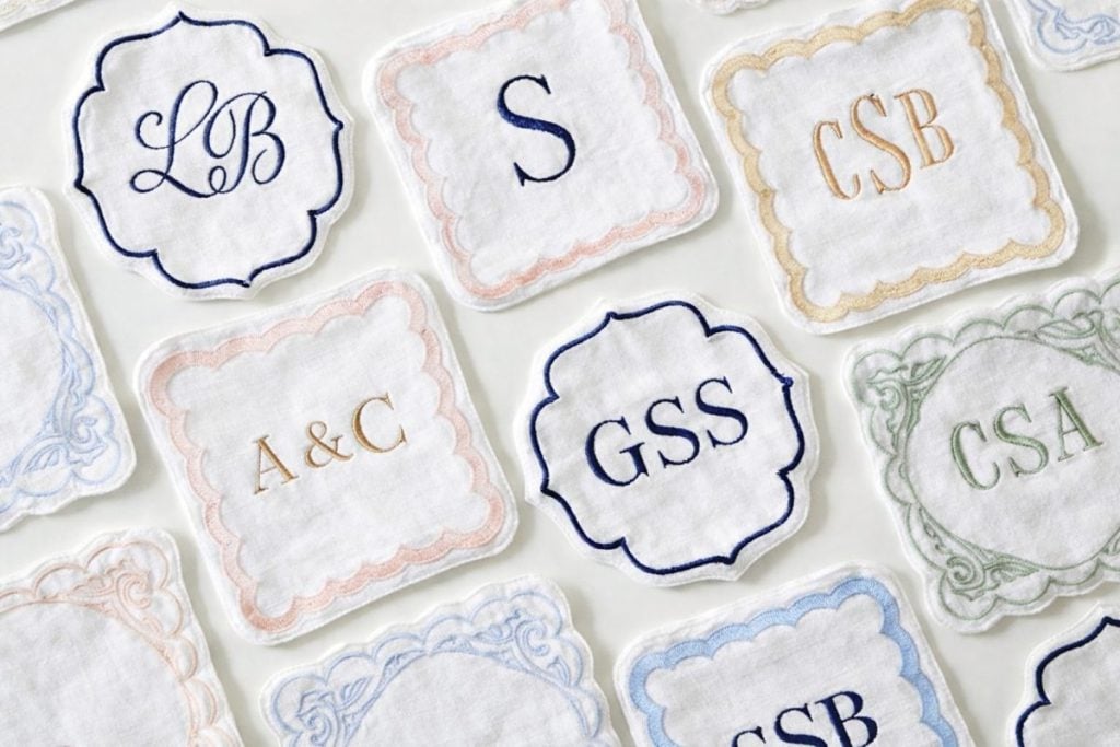 A collection of embroidered white fabric coasters, each featuring different monogrammed initials in colored thread within decorative borders.