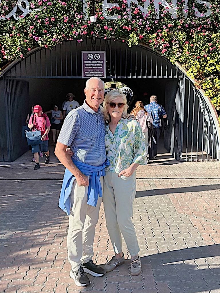 An older couple stands smiling in front of an arched entrance decorated with spring flowers; people are visible in the background on a sunny day.