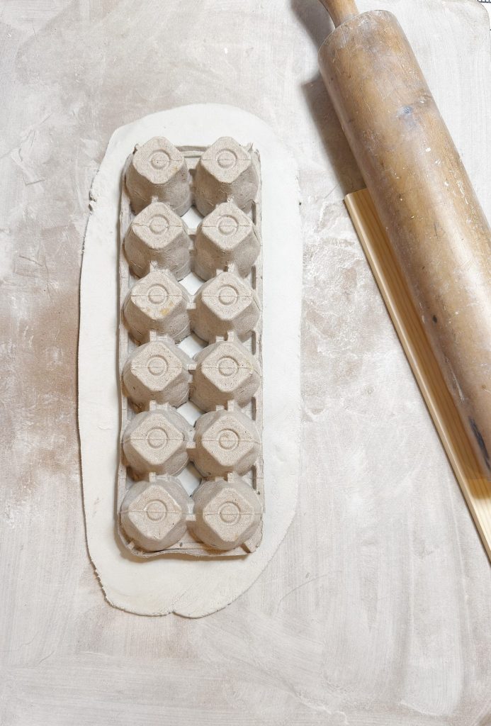 An egg carton is pressed into a sheet of rolled-out dough on a floured surface with a wooden rolling pin nearby.