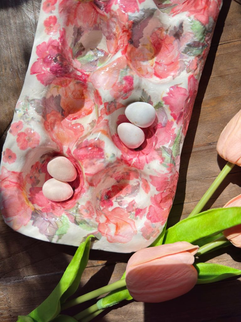A floral-patterned egg tray holds five small pastel-colored eggs, with pink tulips placed nearby on a wooden surface.