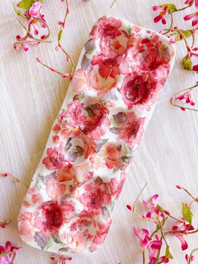 A rectangular ceramic tray with a pink and red floral pattern lies on a light wooden surface, surrounded by pink flower sprigs.