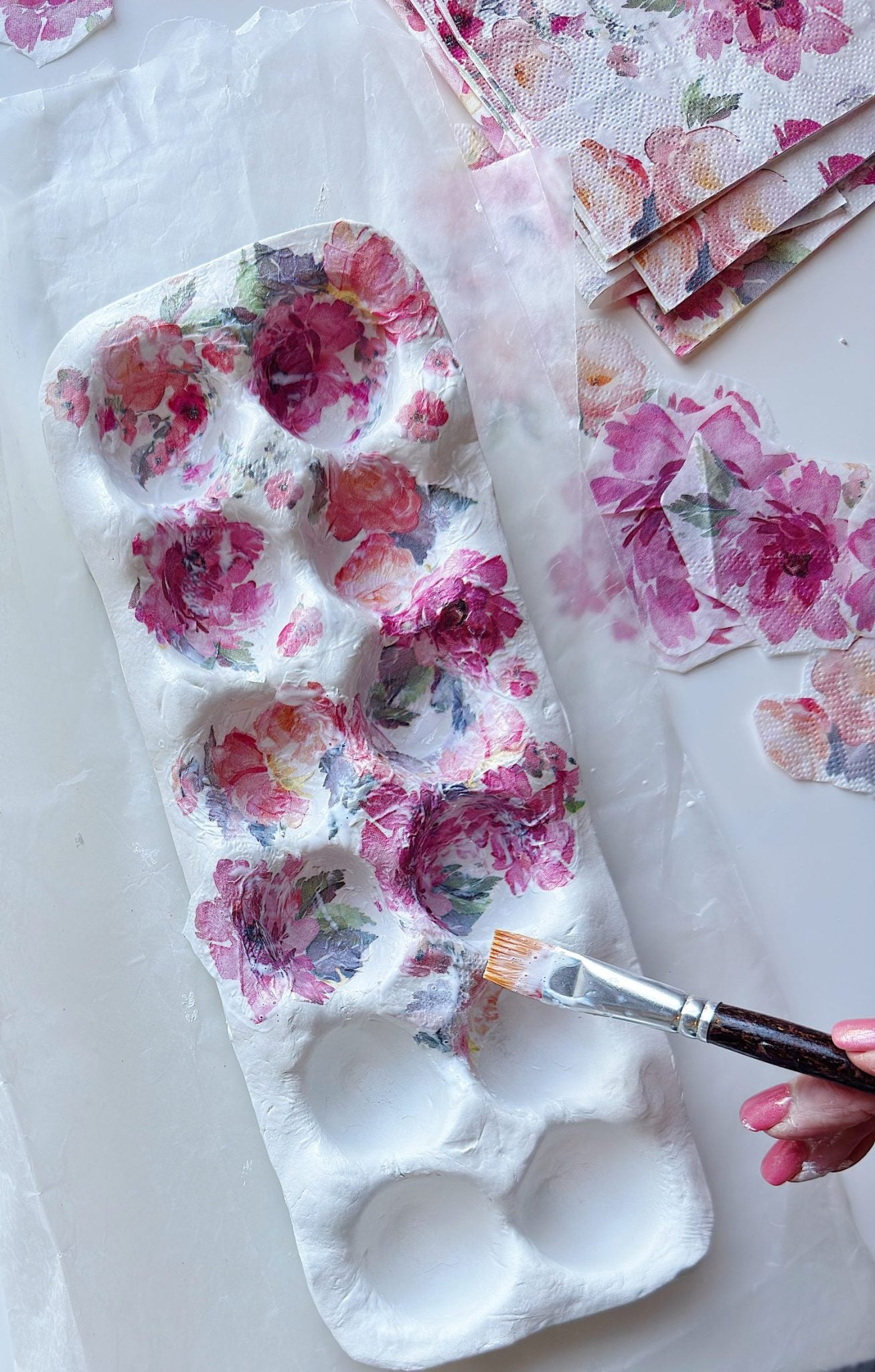 A hand uses a paintbrush to apply floral-patterned paper onto a white ceramic tray with round indentations, with additional floral napkins nearby.