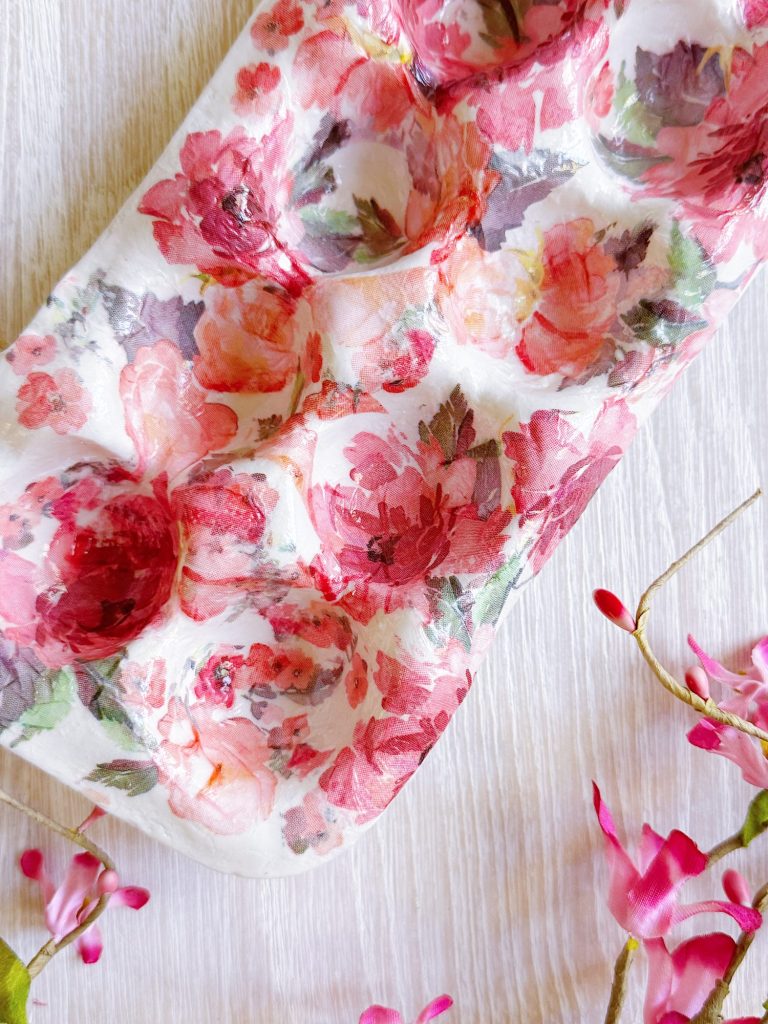 A close-up of a floral-patterned egg carton on a light wooden surface, with pink artificial flowers nearby.