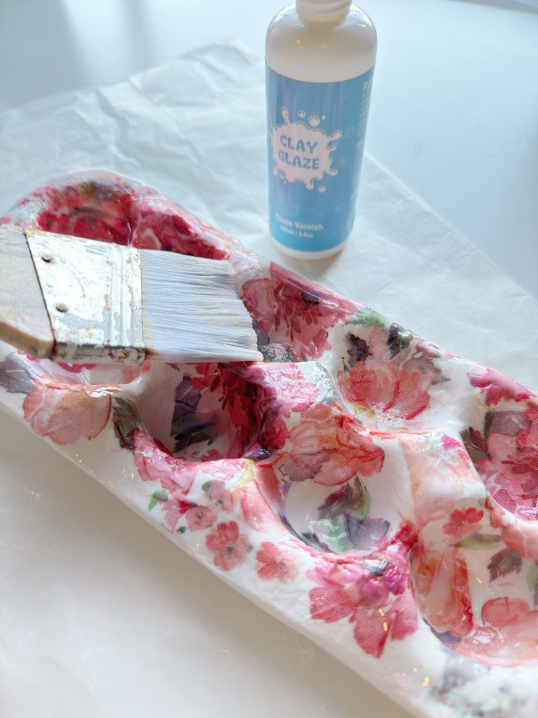 A hand uses a paintbrush to apply clay glaze to a floral-patterned decorative tray; a bottle labeled "Clay Glaze" is nearby.