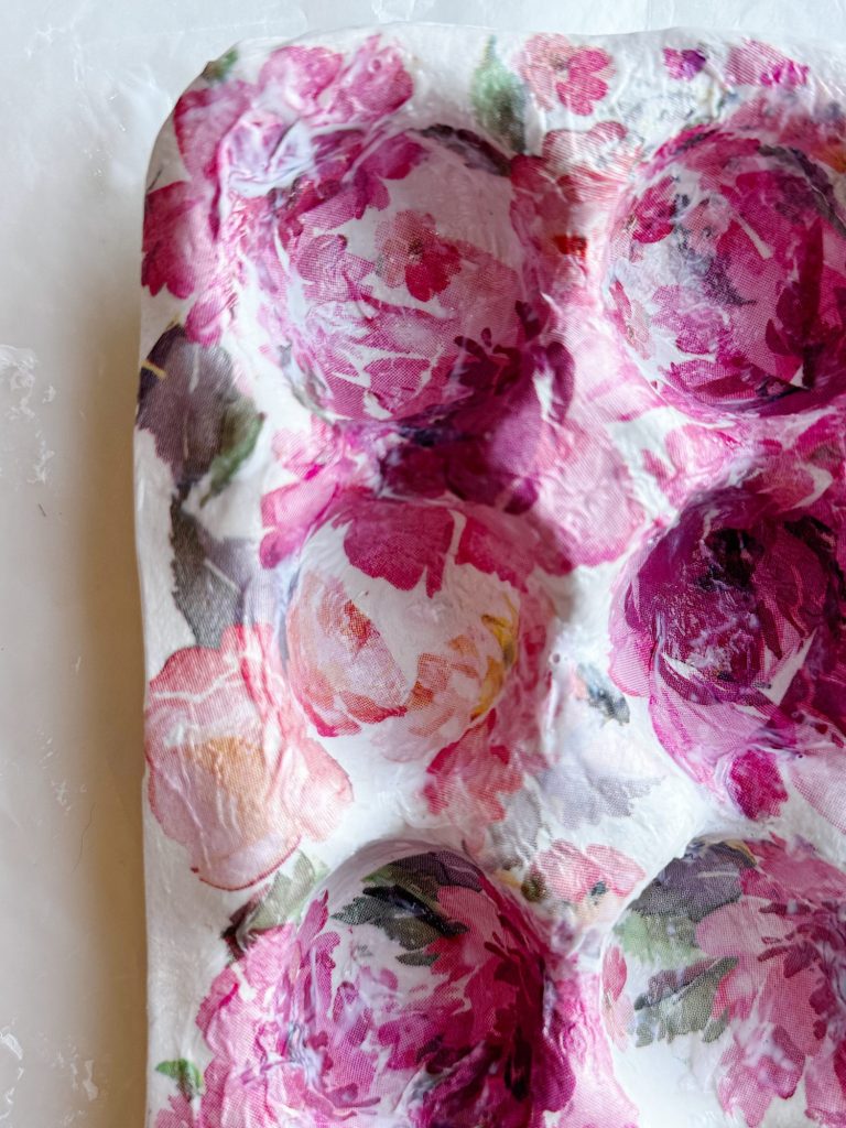 A close-up of a floral-patterned egg carton with shades of pink, purple, and white flowers against a light background.