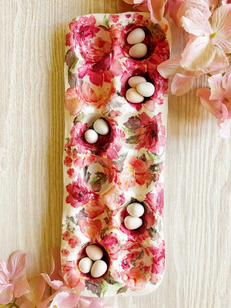 A floral-patterned egg carton holds several small white eggs in some of its compartments, surrounded by pink flowers on a light wooden surface.