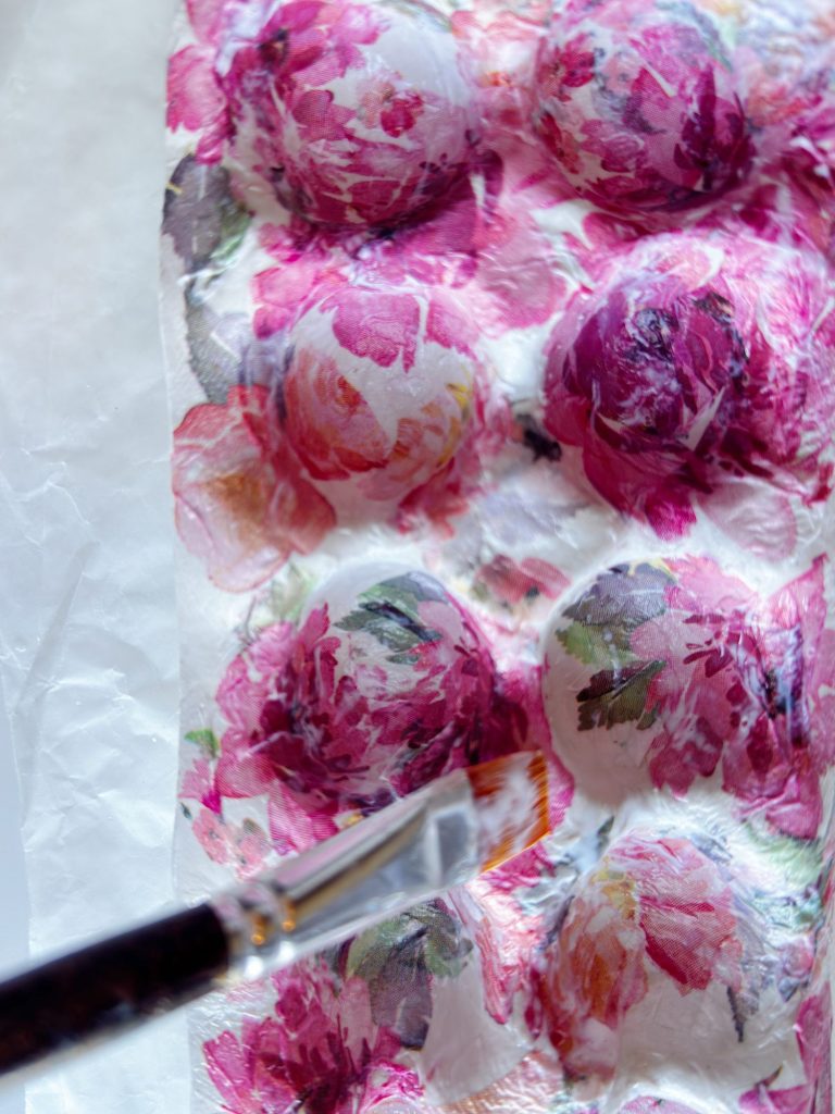 A paintbrush applies glue or paint to a floral-patterned paper napkin covering an egg carton, creating a decoupage art project.