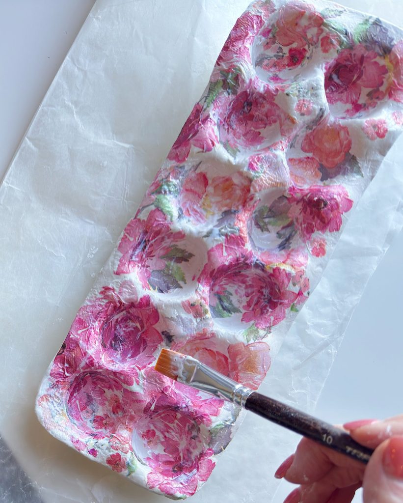 A hand paints a floral pattern onto an egg carton using a paintbrush, with the carton resting on a sheet of wax paper.