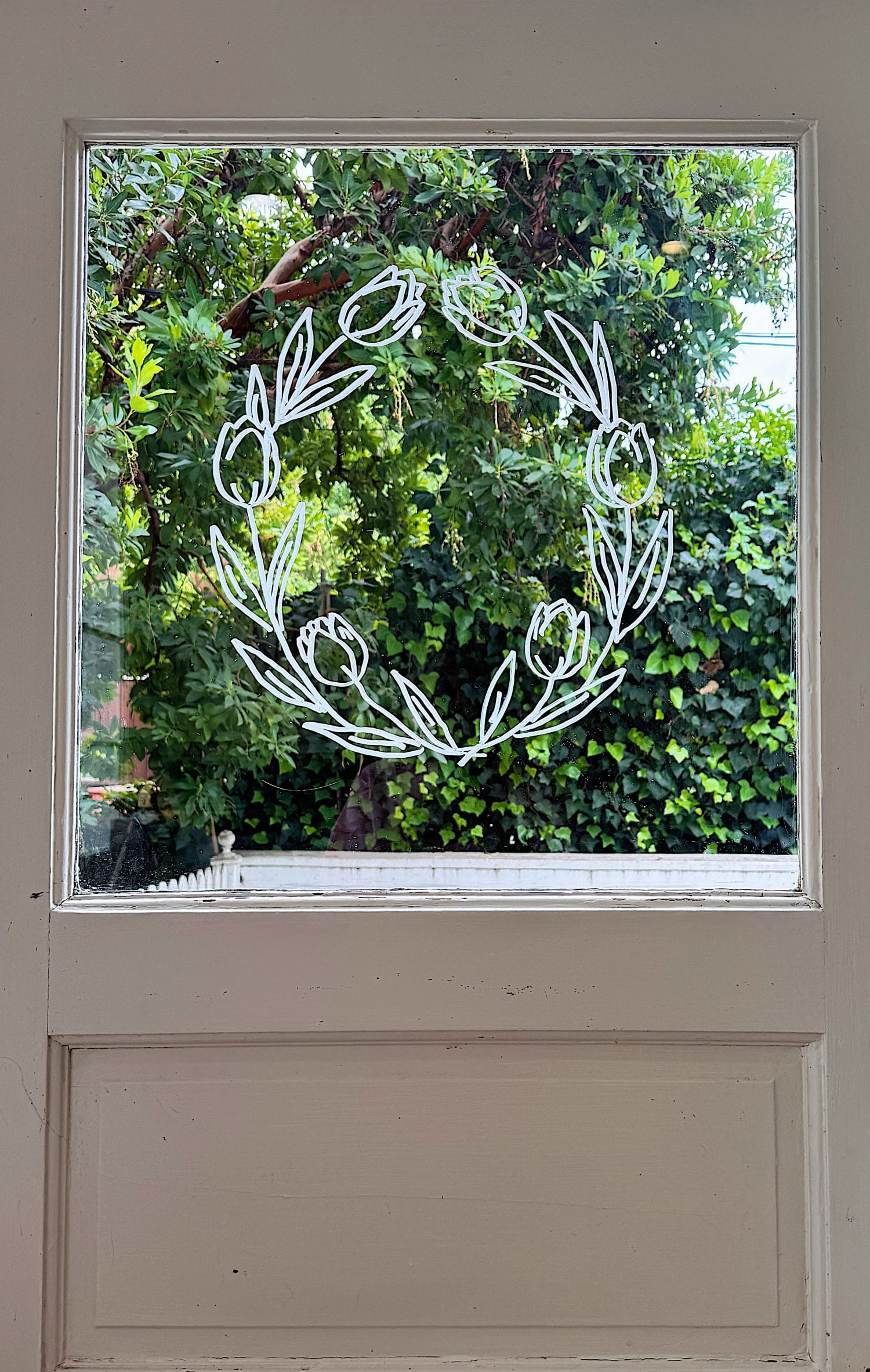 A white floral wreath design is drawn on a window, with green trees and foliage visible outside through the glass.