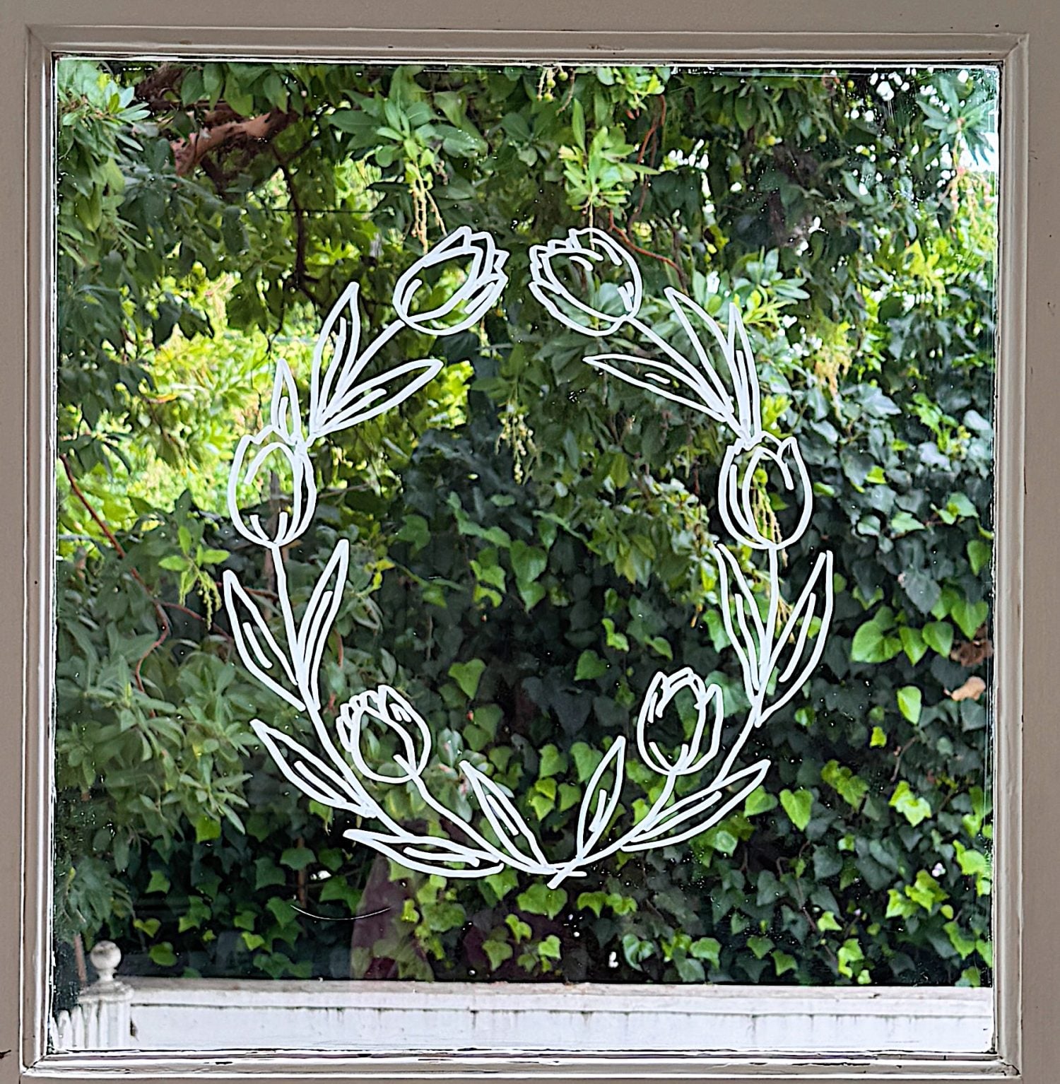 A white floral wreath design is drawn on a window, with green leafy trees and bushes visible outside.