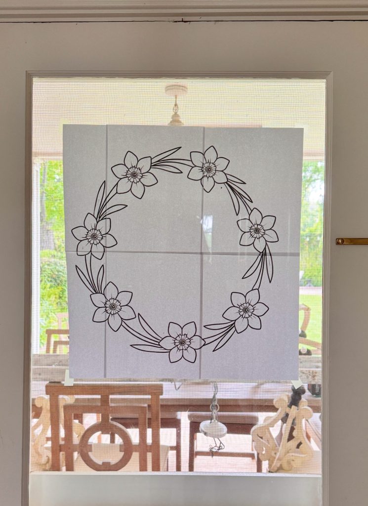 A black-and-white floral wreath design is taped to a glass door, with a porch and outdoor greenery visible through the window.