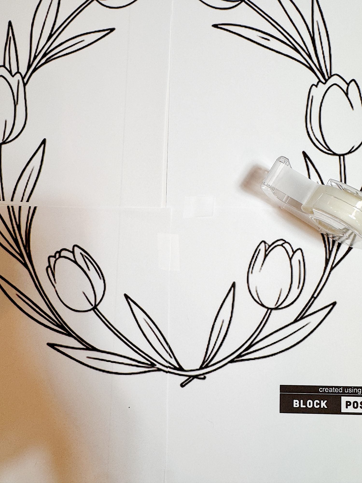 Black and white outline drawing of a floral wreath with tulip-like flowers, assembled from separate papers and taped together; a tape dispenser is placed on the right side.